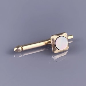 May include: A gold-coloured collar pin featuring a square head with a round, iridescent mother-of-pearl centre. The pin has a long, slender shaft and a pointed tip. The square head measures approximately 0.8 cm across.