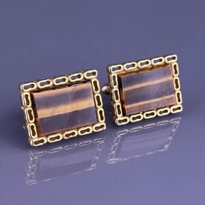 May include: A pair of rectangular cufflinks with a gold-colored frame and a brown, striped stone center. The frame has a repeating linked design. The cufflinks are displayed on a reflective, gray surface.