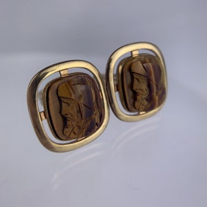 May include: A pair of gold cufflinks with square frames and brown tiger's eye stone insets. The stone insets feature carved faces.