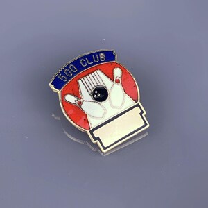 May include: A gold-colored pin with a red, white, and blue design. The pin features a bowling ball and three bowling pins. The text "500 CLUB" is on the top of the pin.