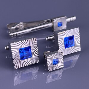 Könnte beinhalten: Ein silberfarbenes formelles Set mit einer Krawattenklammer und Manschettenknöpfen, jeweils mit einem quadratischen, strahlenden Design. Die Mitte jedes Stücks zeigt einen leuchtend blauen quadratischen Edelstein. Das Set enthält zwei Manschettenknöpfe und einen Hemdknopf.