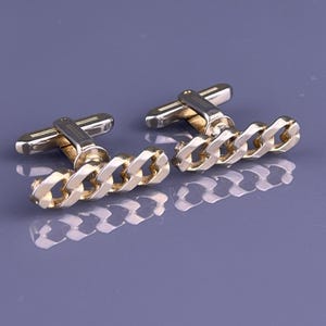 May include: A pair of gold-toned cufflinks with a chain link design. The cufflinks have a rectangular bar and a chain of interlocking links. The background is a solid, muted purple.