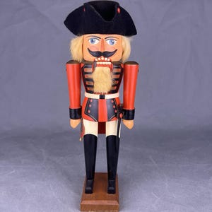 Puede incluir: Figura de cascanueces de madera con forma de soldado. La figura tiene un sombrero negro, pelo rubio, bigote y barba de piel sintética. Viste un uniforme rojo y negro con botas negras y está sobre una base de madera.