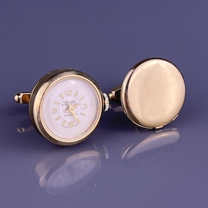 May include: Gold-toned watch cufflinks with a white watch face. The watch face has black numerals and hands, with the brand name "Brevfield" printed on the face. The cufflinks are round and have a classic design.