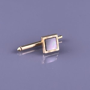 May include: A gold tie tack featuring a square mother-of-pearl inlay. The square is framed by a textured gold border. The tack has a long, slender post with a pointed tip.