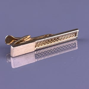 May include: A gold-toned tie clip with a rectangular design. The clip features a decorative filigree pattern and a rectangular clasp. The tie clip is designed to secure a tie to a shirt.