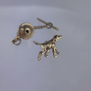Puede incluir: Un broche de corbata dorado con una figura de perro esculpida detallada. El broche de corbata tiene una cadena y una base redonda texturizada.