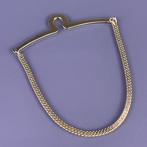 May include: A gold-toned tie chain with a curved bar and a chain that drapes in a U-shape. The chain is made of small, interwoven links. The background is a solid, muted purple.