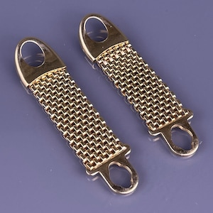 May include: Two gold-colored metal mesh straps with oval loops at the top and open teardrop-shaped loops at the bottom. The straps are made of a tightly woven mesh pattern, and the metal has a shiny finish. The background is a solid, muted purple.