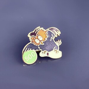 May include: A gold-colored pin featuring a cartoon owl wearing a white shoe and holding a green bowling ball with the number 500 on it.