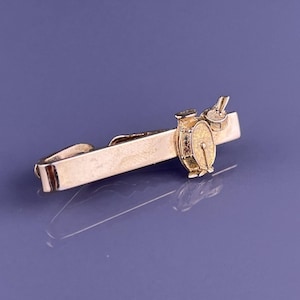 Könnte beinhalten: Eine goldfarbene Krawattenklammer mit einem detaillierten Schlagzeug-Design. Der Stab hat eine rechteckige Form mit einem Verschluss und einem Schlagzeug-Charm. Das Schlagzeug-Set umfasst eine Trommel, ein Becken und Trommelstöcke.