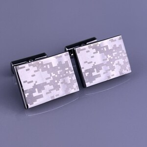 May include: A pair of rectangular cufflinks with a digital camouflage pattern in shades of gray and white. The cufflinks have a shiny, silver-colored metal frame and a black backing.