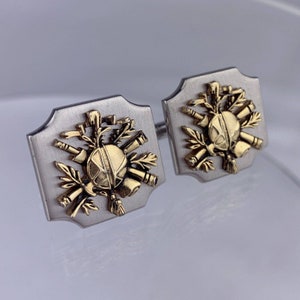 Puede incluir: Un par de gemelos de plata con detalles dorados. Los gemelos presentan un diseño de un escudo rodeado de varias herramientas y ramas.