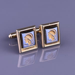 May include: A pair of gold-toned cufflinks with a rectangular design. Each cufflink features a light blue square with a gold-colored profile of a person wearing a helmet. The cufflinks have a classic, elegant style.