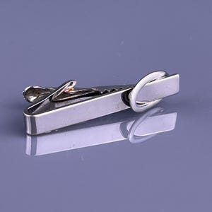 Può includere: Un fermacravatta argentato con un corpo rettangolare e un design curvo. La clip ha un bordo strutturato e un piccolo elemento decorativo. Il fermacravatta è mostrato su una superficie riflettente. Misura circa 5 cm.