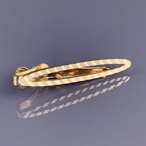 Könnte beinhalten: Eine goldfarbene Krawattenklammer mit einem strukturierten, gestreiften Design. Die Klammer hat eine rechteckige Form mit abgerundeten Enden und einem Klappverschluss. Die Klammer dient dazu, eine Krawatte zu halten.