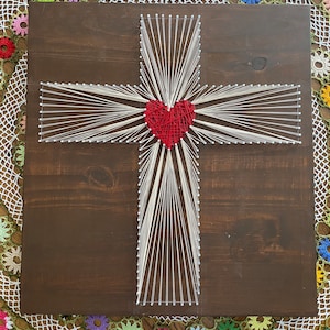 Puede incluir: Una cruz de madera con un corazón rojo en el centro, creada con arte de hilo. La cruz está rodeada de hilo blanco, y el corazón está hecho de hilo rojo. El fondo es de madera marrón oscura.