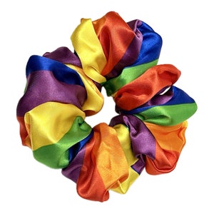 May include: A circular hair scrunchie made of smooth, shiny fabric featuring a repeating pattern of vibrant rainbow stripes in red, orange, yellow, green, blue, and purple. The accessory has a gathered, ruffled texture throughout its circumference.