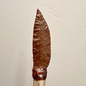 May include: A brown stone knife blade mounted on a bone handle. The blade is pointed and has a rough, textured surface.