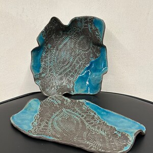 Puede incluir: Dos platos de cerámica con un esmalte azul y un patrón de encaje negro. Los platos tienen una forma irregular y un aspecto rústico y hecho a mano.
