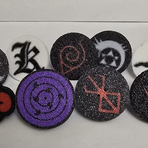 Puede incluir: Una colección de botones redondos y texturizados con varios diseños. Los botones presentan símbolos y gráficos en negro, blanco, rojo y morado. Un botón tiene forma de nube roja.