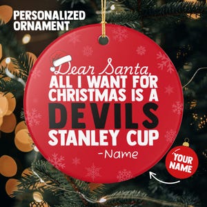 May include: A red Christmas ornament with the text "Dear Santa, ALL I WANT FOR CHRISTMAS IS A DEVILS STANLEY CUP -Name". The ornament has a Santa hat graphic and snowflake designs. A smaller ornament says "YOUR NAME".