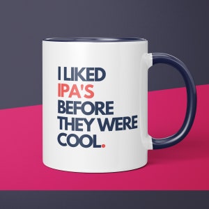 May include: White ceramic coffee mug with a navy blue rim and handle. The mug has the text "I LIKED IPA'S BEFORE THEY WERE COOL." printed in navy blue and red.