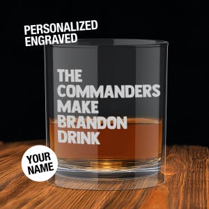 May include: A personalized engraved whiskey glass with the text "THE COMMANDERS MAKE BRANDON DRINK" in white. The glass contains amber liquid and rests on a wooden surface. A white circle with "YOUR NAME" is also visible.