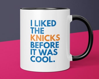 Pode incluir: Caneca de café de cerâmica branca com borda e alça pretas. A caneca tem o texto "I LIKED THE KNICKS BEFORE IT WAS COOL." impresso em azul e laranja.