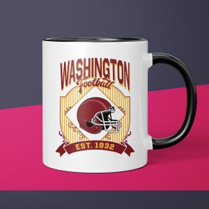 May include: A white ceramic mug with a black handle and interior. The mug features a maroon football helmet graphic and the words "WASHINGTON Football" in a vintage style. The text "EST. 1932" is also displayed.