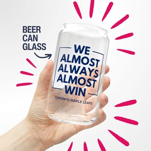 May include: A clear beer can glass with the text "WE ALMOST ALWAYS ALMOST WIN" and "TORONTO MAPLE LEAFS" in blue. The words "BEER CAN GLASS" are to the left of the glass. Pink lines radiate from the glass.