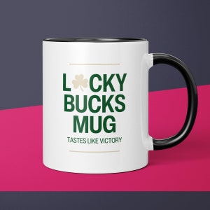 May include: White ceramic mug with a black rim and handle. The mug has green shamrock and the text "Lucky Bucks Mug" in green and gold. The text "Tastes like victory" is in gold.