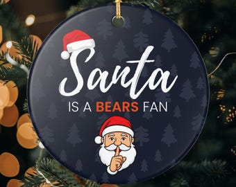 Może przedstawiać: Okrągła ozdoba świąteczna z napisem "Santa is a Bears Fan". Ozdoba przedstawia czapkę Świętego Mikołaja i kreskówkowego Świętego Mikołaja z palcem na ustach. Tło jest granatowe z wzorem małych sosen.