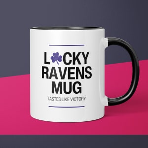 May include: White ceramic mug with a black rim and handle. The mug has a black and white graphic that says "Lucky Ravens Mug" with a four-leaf clover and the text "Tastes like victory."