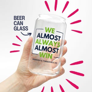 May include: A clear glass beer can with the text "WE ALMOST ALWAYS ALMOST WIN" in green and white, with "MINNESOTA TIMBERWOLVES" below. The words "BEER CAN GLASS" are on the left. The glass is held by a hand, with pink lines radiating from the top.