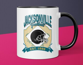 May include: White ceramic mug with a black handle and interior. The mug features a teal and gold design with the words "JACKSONVILLE Football" and a black football helmet. The text "EST. 1995" is also included.