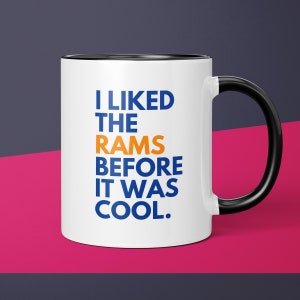 May include: White ceramic coffee mug with a black rim and handle. The mug has the text "I LIKED THE RAMS BEFORE IT WAS COOL." in blue and orange lettering.