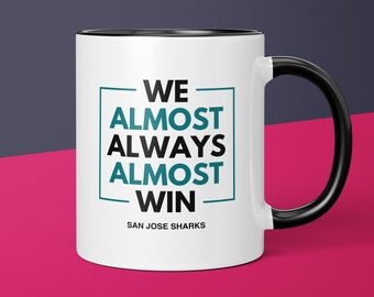 Pode incluir: Caneca de café branca com borda e alça pretas. A caneca tem um gráfico azul-esverdeado e preto que diz "WE ALMOST ALWAYS ALMOST WIN" com as palavras "SAN JOSE SHARKS" abaixo.