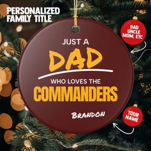 May include: A round, burgundy-coloured ceramic Christmas ornament featuring the white and yellow text "JUST A DAD WHO LOVES THE COMMANDERS" and the name "BRANDON" in white. The ornament includes a gold hanging string and a small hole at the top.