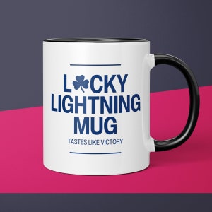 May include: White ceramic mug with a black rim and handle. The mug has blue text that reads "Lucky Lightning Mug" with a shamrock symbol and "Tastes Like Victory" underneath.