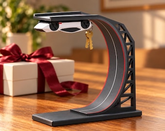 May include: A black key holder shaped like a curved race track ramp with red accents. A white toy sports car is attached to the underside of the top platform, featuring a metal ring that holds a set of gold-colored keys hanging down.