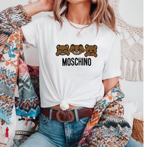 May include: White t-shirt with a graphic of three brown teddy bears covering their eyes, nose, and mouth. The text "MOSCHINO" is printed below the bears.