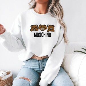 May include: White sweatshirt with a brown bear graphic and the text "MOSCHINO" printed on the front. The bear is covering its eyes, mouth, and ears.