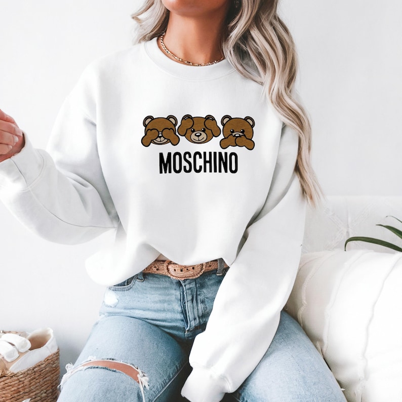 May include: White sweatshirt with a brown bear graphic and the text "MOSCHINO" printed on the front. The bear is covering its eyes, mouth, and ears.