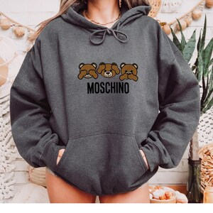 May include: A dark gray hoodie with a black graphic of three brown teddy bears covering their eyes, nose, and mouth. The text "MOSCHINO" is printed below the bears.