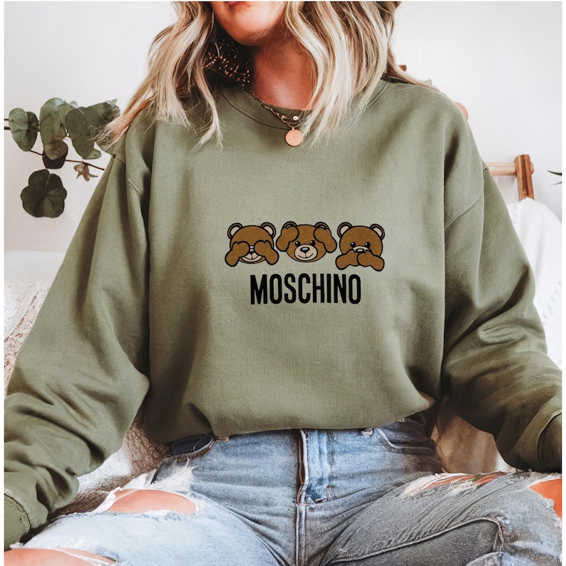 May include: A green sweatshirt with a graphic of three brown bears covering their eyes, mouth, and ears. The text "MOSCHINO" is printed below the bears.