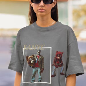 May include: A grey t-shirt featuring a graphic print of Kanye West holding a bear and another bear sitting down. The text "KANYE" is printed above the graphic.