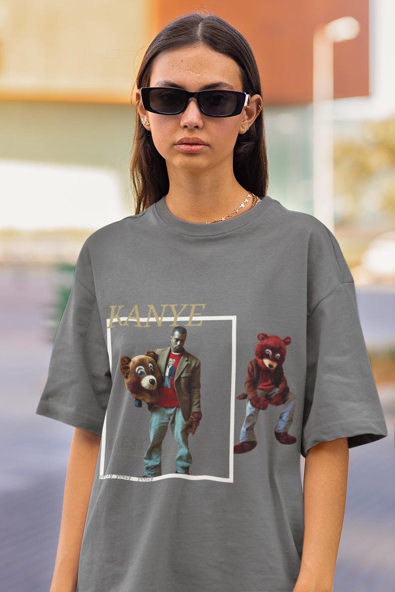 May include: A grey t-shirt featuring a graphic print of Kanye West holding a bear and another bear sitting down. The text "KANYE" is printed above the graphic.
