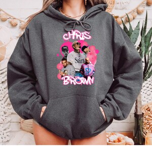 May include: A dark gray hoodie with a pink graphic print of Chris Brown. The graphic features multiple images of Chris Brown in different poses and outfits. The text "Chris Brown" is written in pink letters above the images.