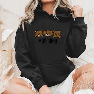 May include: Black hoodie with a graphic of three brown teddy bears covering their eyes, mouth, and ears. The text "MOSCHINO" is printed below the bears.